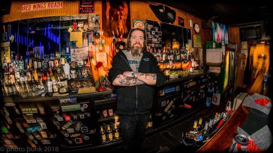 Jimmy Doom stands in front of a full bar of liquor bottles