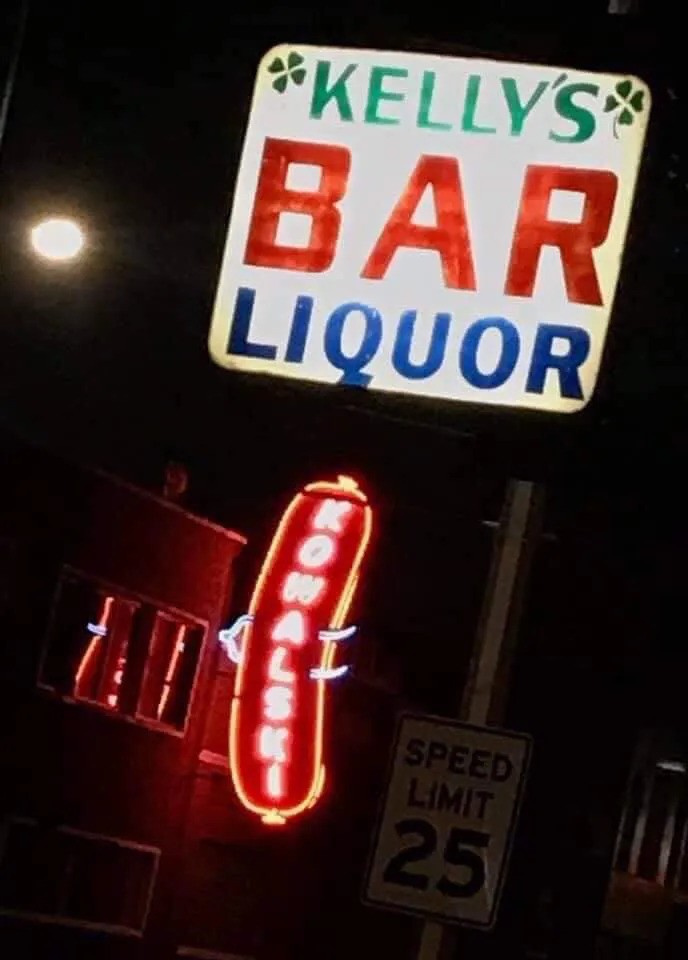 A sign for Kelly’s Bar lights the night, juxtaposed with the neon Kowalski sign in the background